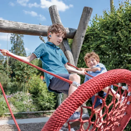 Zwei Jungen klettern auf einem Spielplatz über ein rotes Kletternetz, der ältere trägt ein blaues Hörgerät hinter dem Ohr.