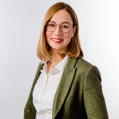 Kristin Nartschik im grünen Blazer über weißer Bluse vor hellem Hintergrund, sie trägt eine runde Brille und lächelt in die Kamera.